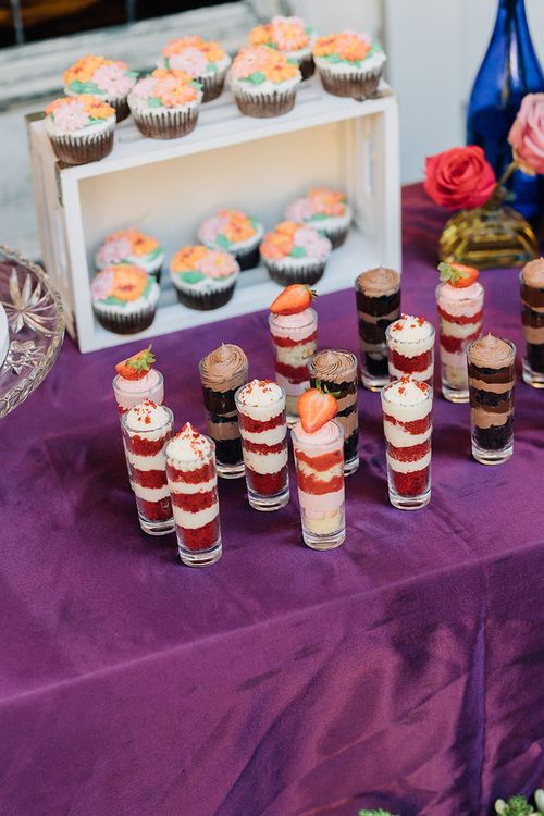 A dessert table with cupcakes and layered shots in glass cups on a purple tablecloth.