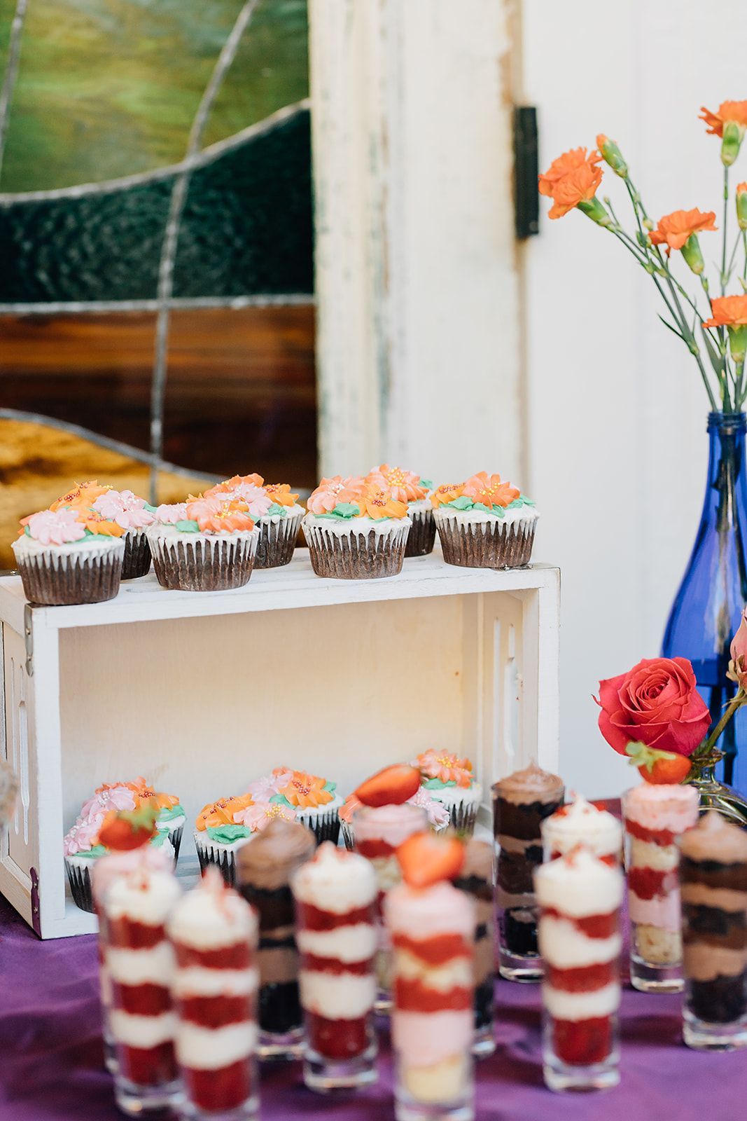 Cupcakes and layered desserts on a table with flower arrangements. Baked goods have orange frosting and strawberries.