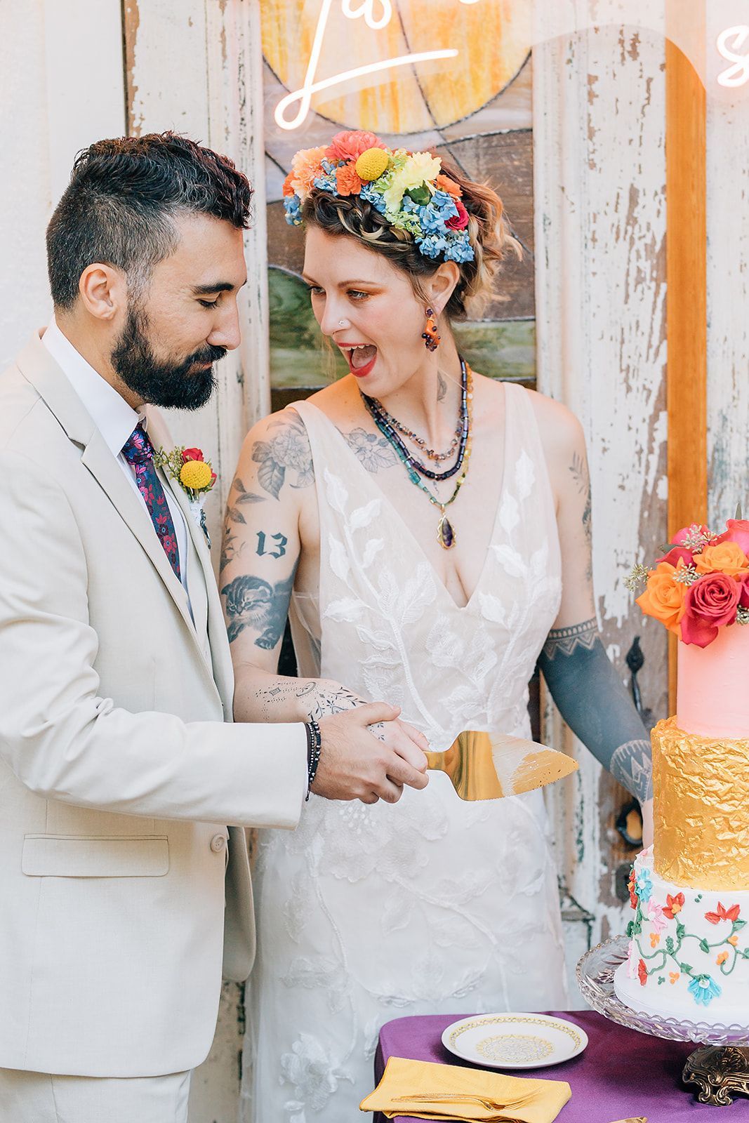 A bride and groom cut their wedding cake. The bride wears a flower crown, tattoos, and a surprised expression; the groom wears a suit.