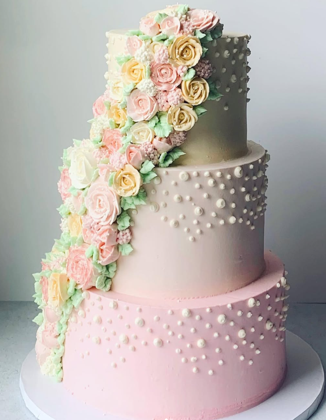 Three-tiered cake with cascading buttercream flowers. The bottom tier is pink, the middle is light pink with pearl dots, and the top is off-white.