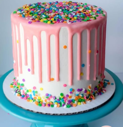 Pink drip cake with colorful sprinkles on top and around the base, sitting on a blue cake stand.