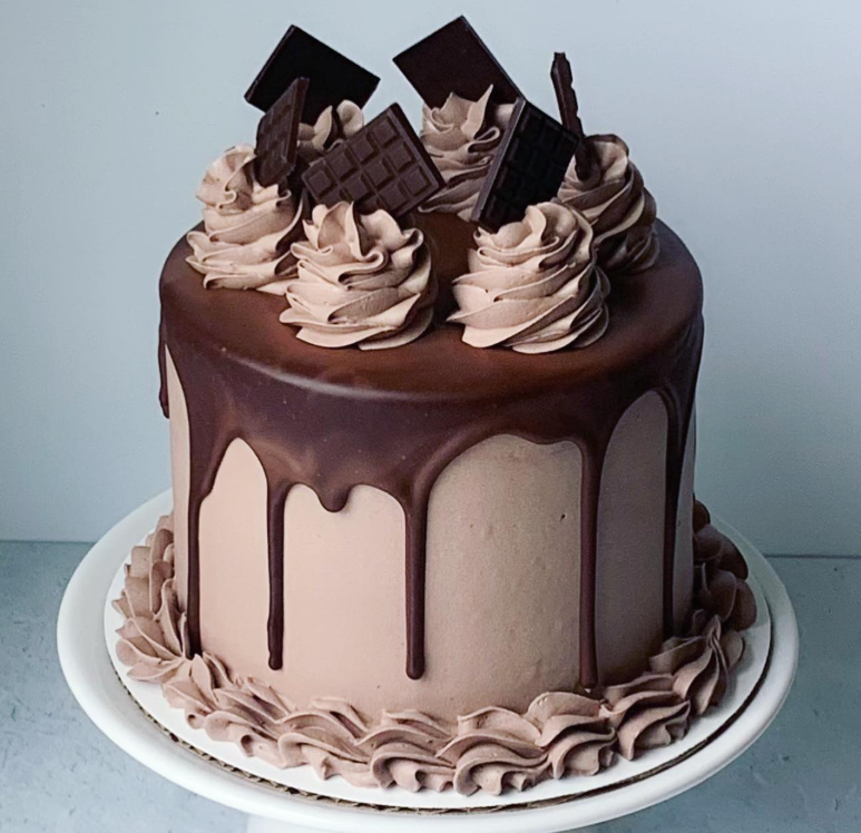Chocolate cake with chocolate ganache drip, topped with whipped cream rosettes and chocolate squares on a white cake stand.