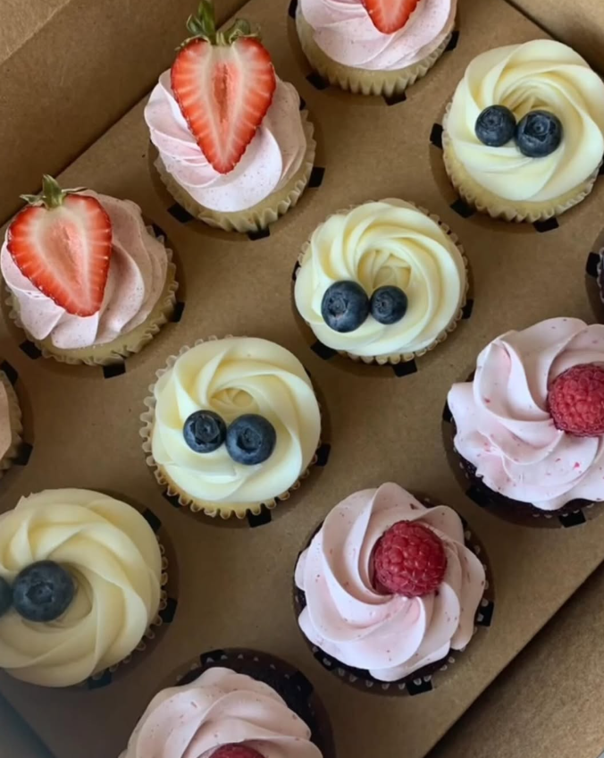 Cupcakes in a box: various flavors with pink and white frosting, topped with strawberries, raspberries, and blueberries.