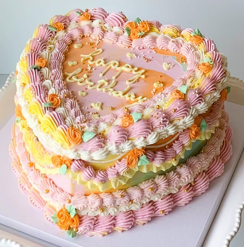 Heart-shaped cake with three tiers, decorated with pink, yellow, and orange buttercream, and
