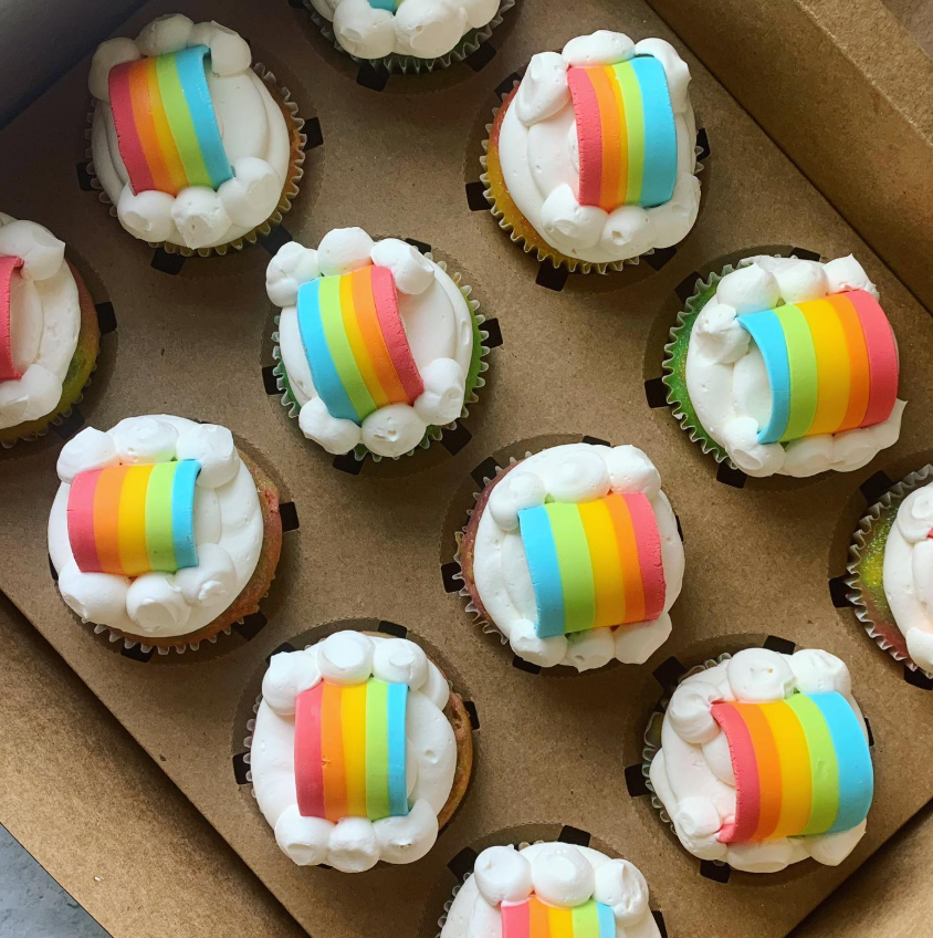 Box of cupcakes topped with white frosting and rainbow-colored fondant.