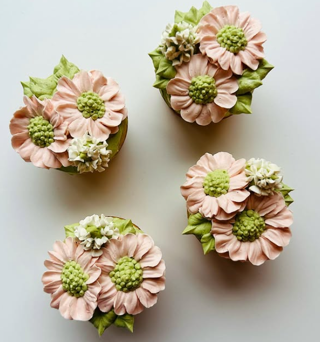 Four cupcakes decorated with light pink buttercream flowers, green centers, and green leaves, arranged on a white surface.