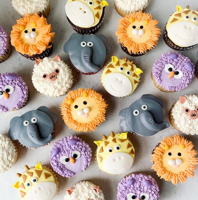 Animal-themed cupcakes with lion, elephant, giraffe, owl, and sheep designs, decorated with frosting and edible details.
