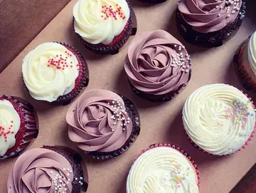 Cupcakes with white and mauve frosting, some sprinkled, arranged in a box.