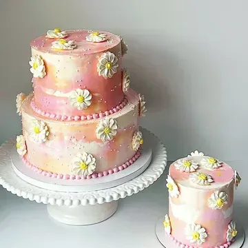 Two tiered pink ombre cake with daisy decorations, next to a smaller matching cake.