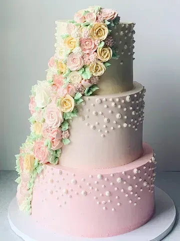 Three-tiered pastel pink cake decorated with piped buttercream flowers and white pearl-like details.