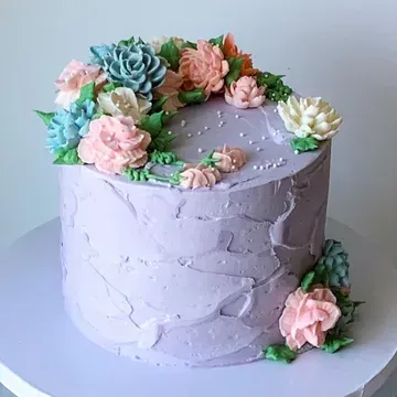 A lavender-frosted cake decorated with pastel-colored buttercream flowers and a textured surface, set on a white surface.