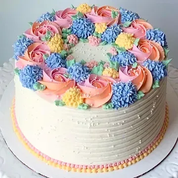 Round cake decorated with pastel-colored buttercream flowers: pink roses, blue and yellow clusters. White frosting with textured sides and a beaded bottom.