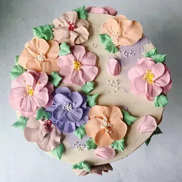 A round cake decorated with buttercream flowers in pastel pink, peach, and purple, with green leaves.
