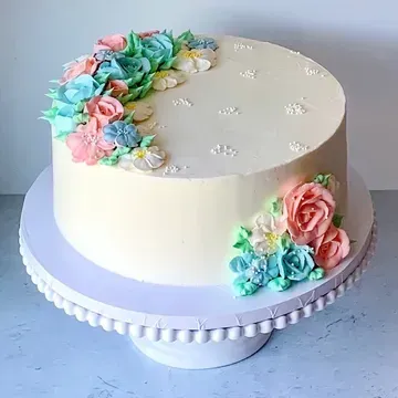 A round cake on a white pedestal decorated with pastel buttercream flowers. The cake has a smooth, cream-colored frosting base.