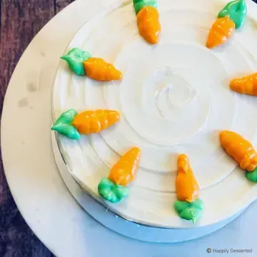A round carrot cake decorated with orange frosting carrots and green frosting leaves on white frosting swirls.