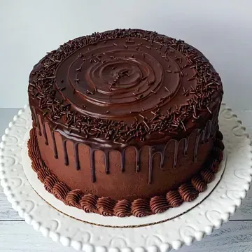 Chocolate cake on a white, scalloped-edge cake stand. The frosted cake has chocolate drips, sprinkles, and a swirl design on top.