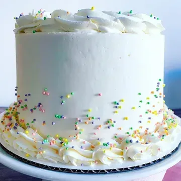 White cake with frosting, sprinkles, and decorative frosting swirls on a white cake stand.