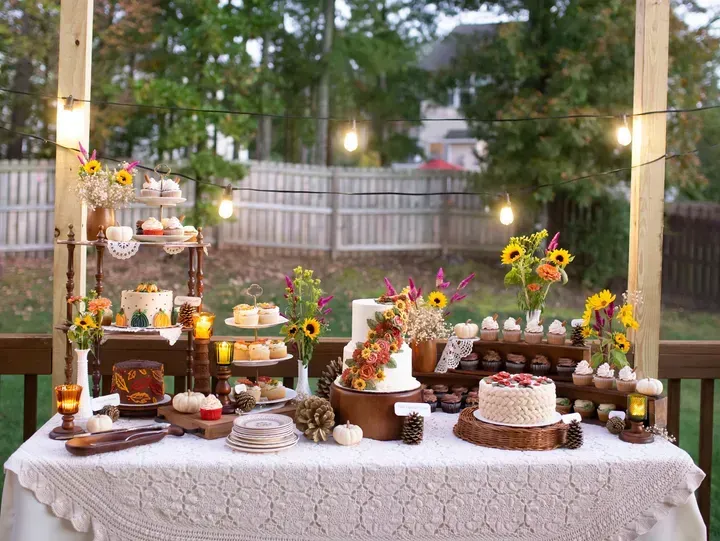 A dessert table on a deck, decorated for fall with cakes, cupcakes, sunflowers, and string lights.