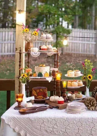 Fall dessert table set on a porch with cakes, cupcakes, pumpkins, flowers, and string lights.