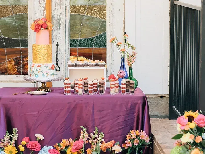 A dessert table with a tiered cake, cupcakes, and parfaits, decorated with flowers, set outdoors with a colorful wall.