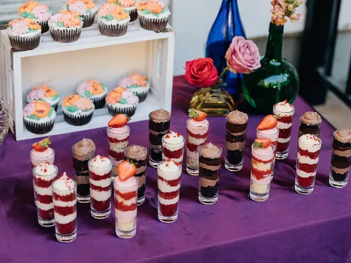 Cupcakes and layered desserts in shot glasses arranged on a purple table; flowers in vases are in the background.