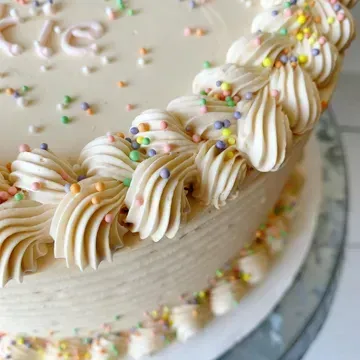 Close-up of a beige frosted cake with piped frosting swirls, pastel sprinkles, and the name