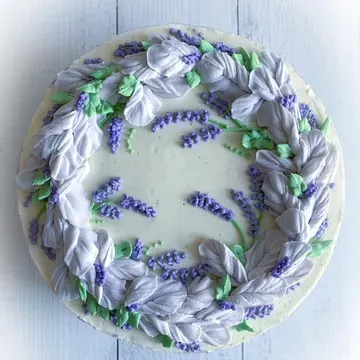 A round cake decorated with a lavender floral wreath made of purple and white frosting, with green leaves.