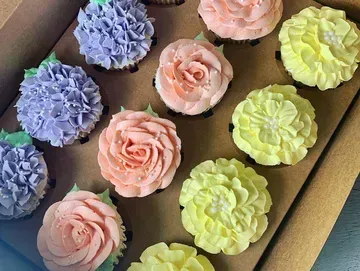 Box of decorated cupcakes with floral frosting in pastel colors: purple, peach, and yellow.