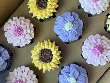 Close-up of a box of flower-shaped cupcakes with yellow, purple, and pink frosting, brown sprinkles in the centers.