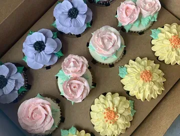 A dozen cupcakes in a box, decorated with pastel buttercream flowers: anemones, roses, and sunflowers.