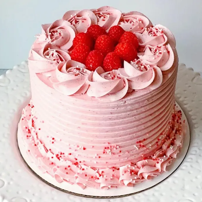 Pink frosted cake decorated with raspberries and sprinkles on a white plate.