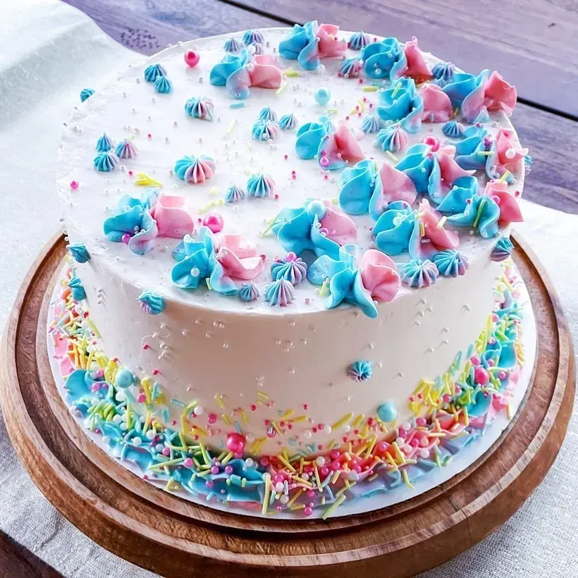 White frosted cake decorated with pink and blue frosting swirls and colorful sprinkles, resting on a wooden cake stand.