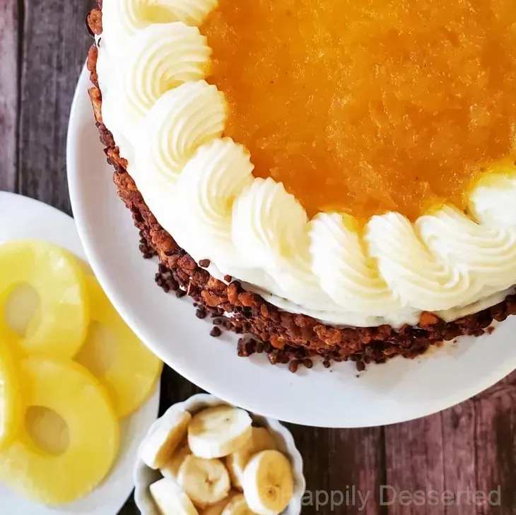 Pineapple cake with whipped cream border and glazed top, beside pineapple slices and bananas.