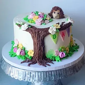 A decorated cake with a woodland theme. It has a tree, flowers, a hedgehog, and a banner that says