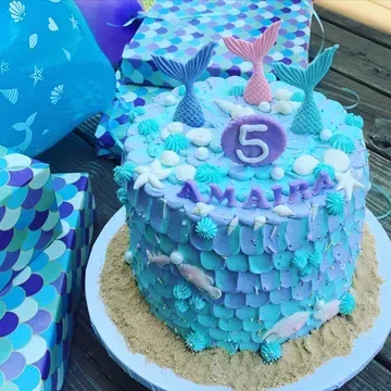 Mermaid-themed cake with blue and purple scale frosting, decorated with mermaid tails, shells, and the name