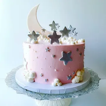 Pink cake decorated with a crescent moon, silver stars, and white cloud-like frosting, on a decorative glass cake stand.