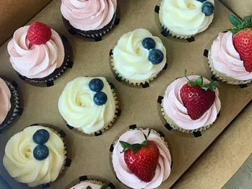 Cupcakes in a box, some with pink frosting and strawberries, others with white frosting and blueberries.