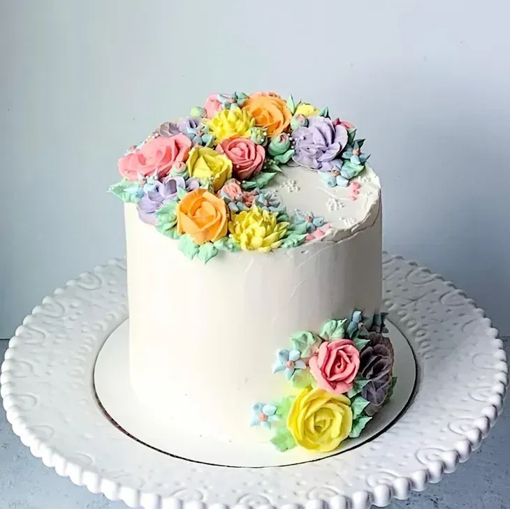 A white cake decorated with pastel buttercream flowers on a white, ornate cake stand.