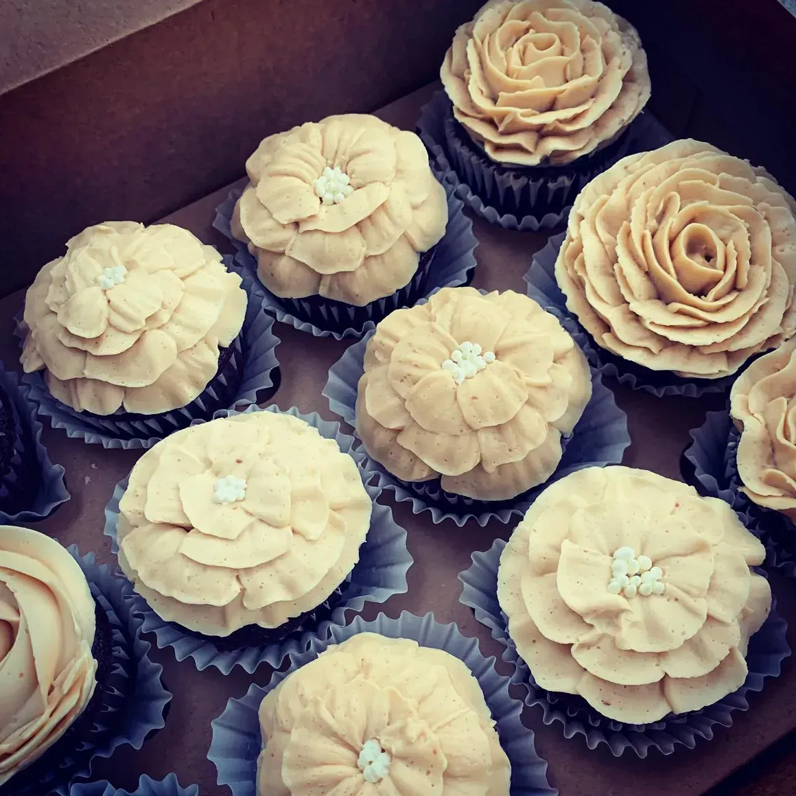 Cupcakes in a box with beige frosting flowers. Some have rosette designs, others are layered petals.
