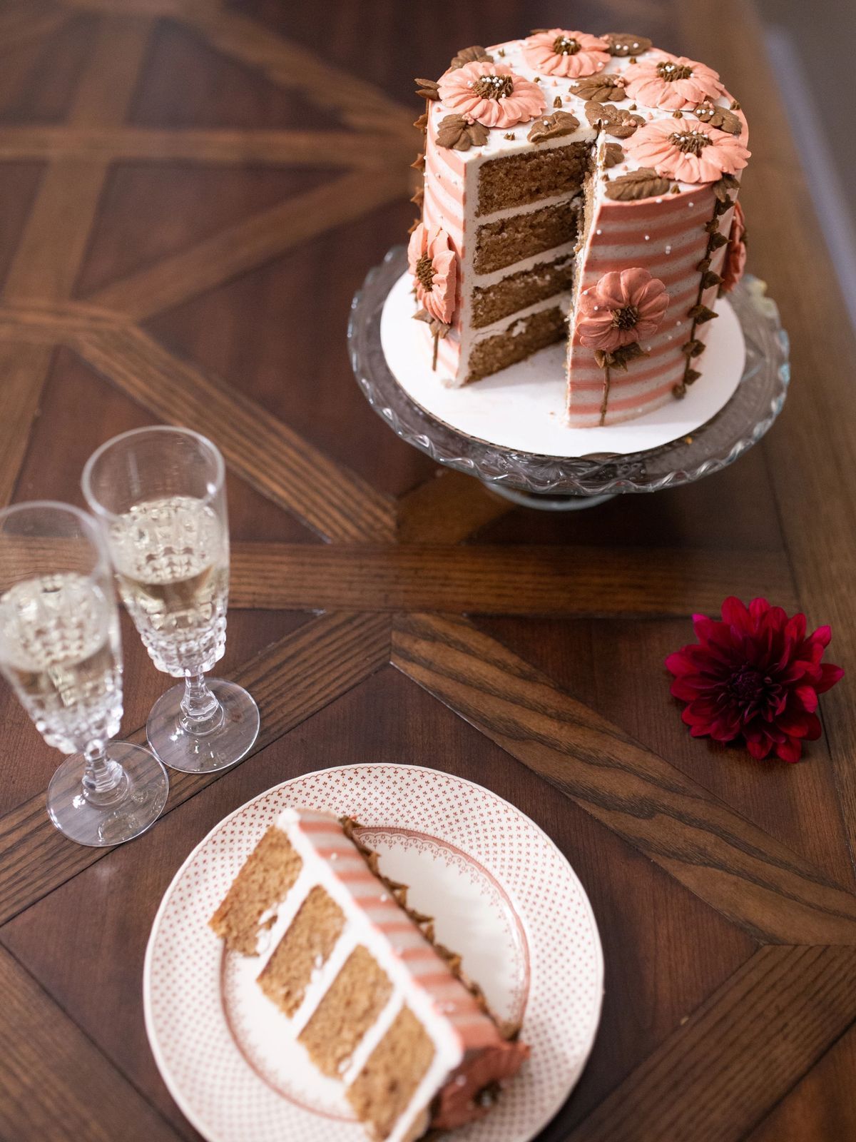 Cake with pink and white stripes, a slice on a plate, champagne glasses, and a flower on a wooden table.