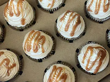 Cupcakes in a box, topped with swirled light-colored frosting and drizzled caramel.