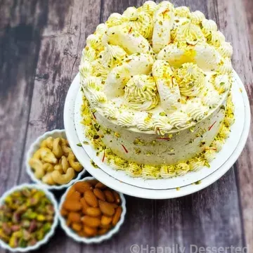 A layered cake decorated with cream frosting and garnished with nuts and saffron, next to bowls of cashews, pistachios, and almonds.