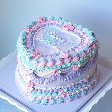 Heart-shaped cake with pastel-colored frosting, including pink, blue, and purple. Decorated with floral accents and piped swirls on a white tray.