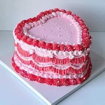 Heart-shaped cake with layers of red and pink frosting, adorned with decorative piping and edible pearls.