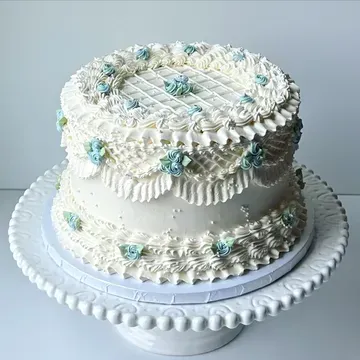Two-tiered white cake with intricate buttercream frosting, decorated with blue floral accents, on a white cake stand.
