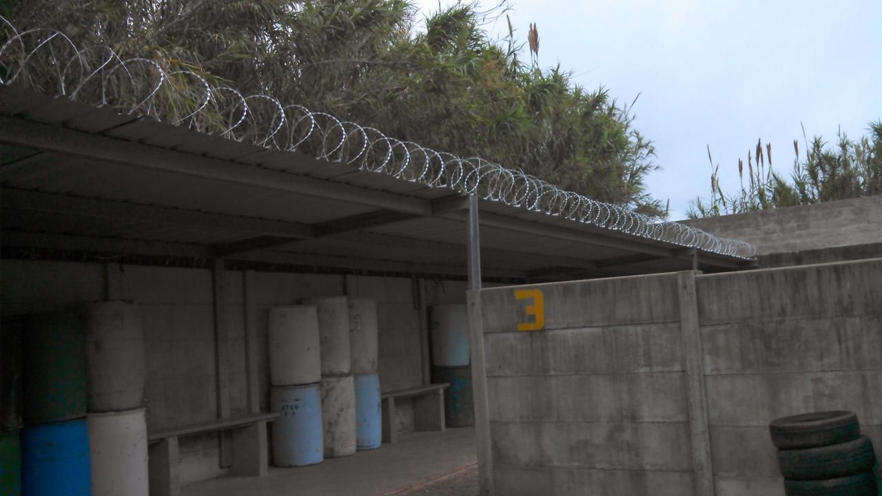 Alambre de púas sobre una pared de hormigón y un techo de metal corrugado, al lado de barriles azules y verdes apilados.