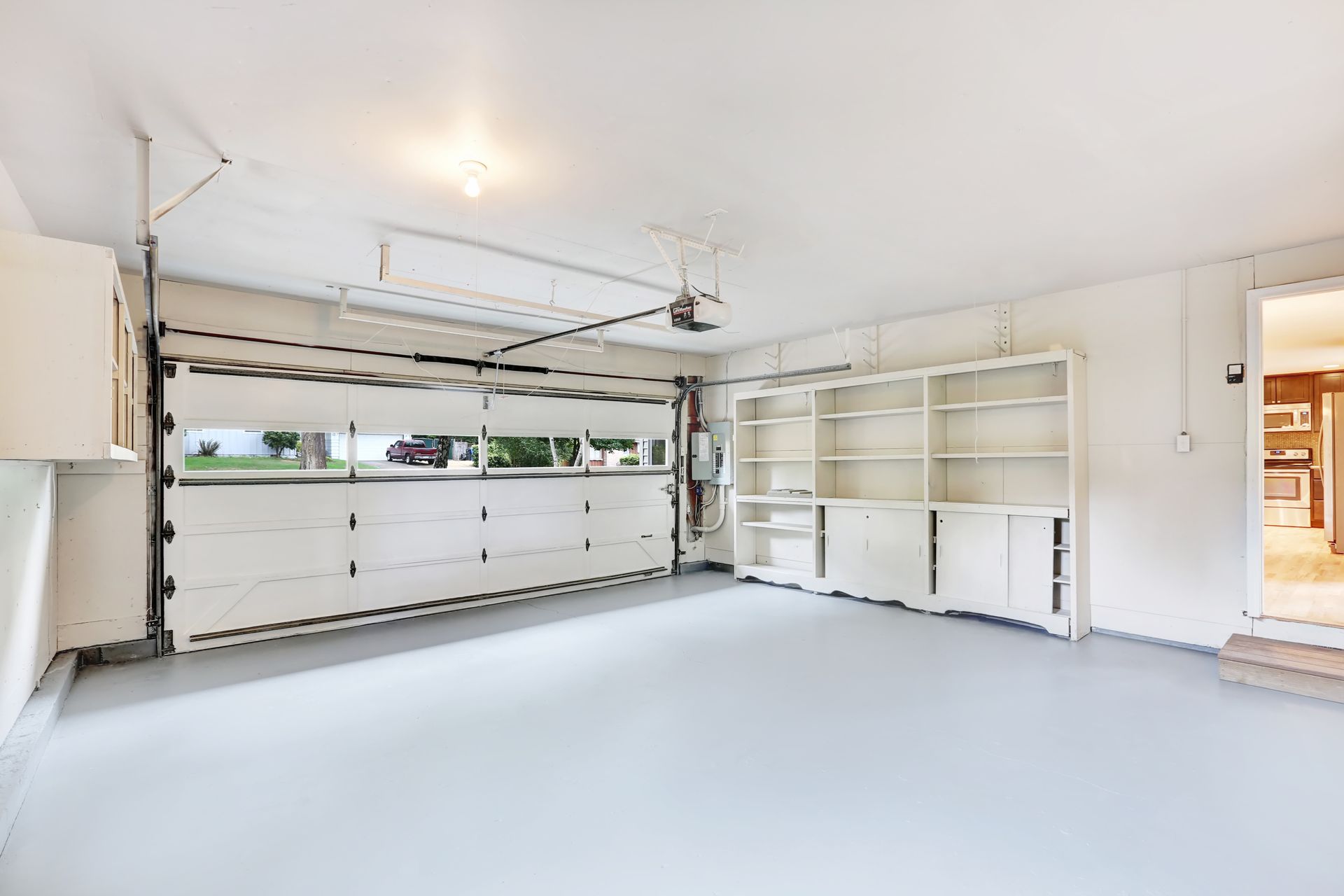 An empty, well-lit garage featuring a white overhead door, light gray floor, and built-in shelving along the side wall.