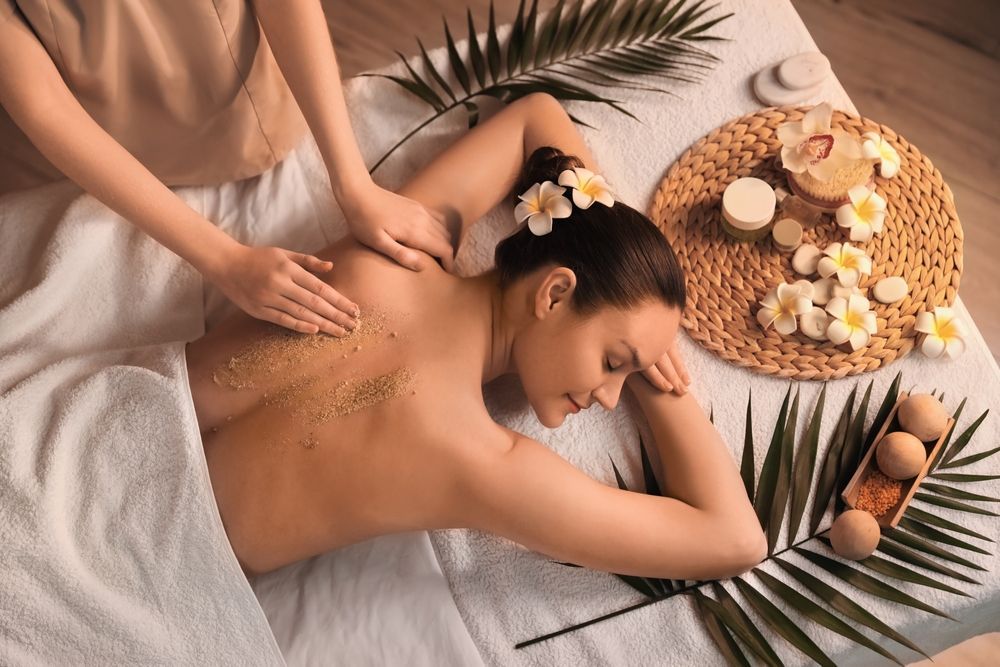 Person receiving back scrub at spa. Palm leaves, flowers, and spa products are visible.