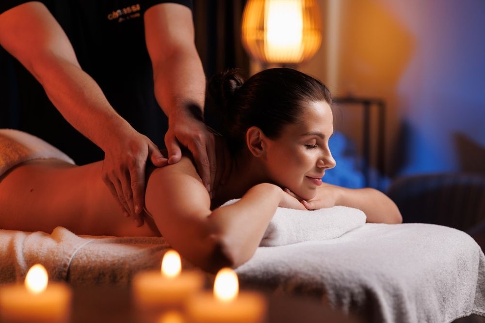 Woman receiving a massage, lying face down on a massage table. Candles glow; a person massages her back.