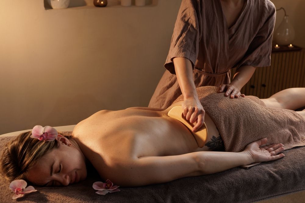 Person receiving back massage in spa. Therapist using wooden tool. Beige setting, floral accents.
